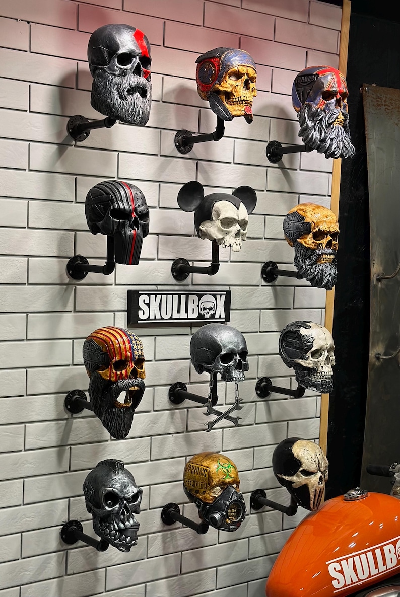 May include: A wall display featuring 12 skull-shaped figurines, each with a unique design and colour scheme. The skulls are mounted on black metal pipes and arranged in a grid pattern. The word "SKULLBOX" is displayed on a black sign in the centre of the wall.