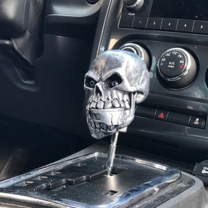 May include: A silver skull gear shift knob for a car. The skull has a menacing expression with black eyes and a toothy grin.