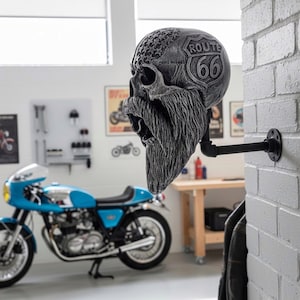 May include: A gray skull-shaped wall hook with a long, textured beard, Route 66 logo, and star pattern. The hook is mounted on a black pipe against a white brick wall. A blue motorcycle is in the background.