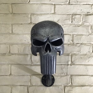 May include: A silver skull wall hook with a black metal pipe attached to a white brick wall.