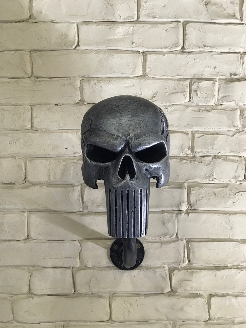 May include: A silver skull wall hook with a black metal pipe attached to a white brick wall.