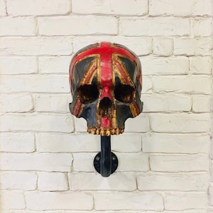 Puede incluir: Un cráneo negro y rojo con un diseño de Union Jack pintado en él. El cráneo está montado en un gancho de metal negro contra una pared de ladrillo blanco.