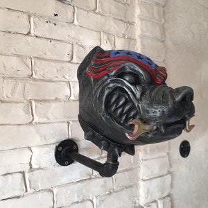 Op de afbeelding: Een zwart en grijs hondenkopbeeld met een rood, wit en blauw bandana. Het beeld is gemonteerd op een zwarte metalen pijp en beugel. De hond houdt twee moersleutels in zijn bek.