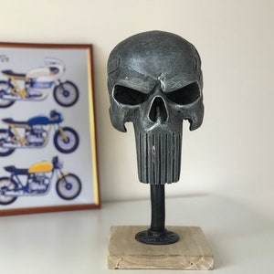 May include: A grey metal skull sculpture with a detailed face and a textured beard. The skull is mounted on a black metal stand with a wooden base.