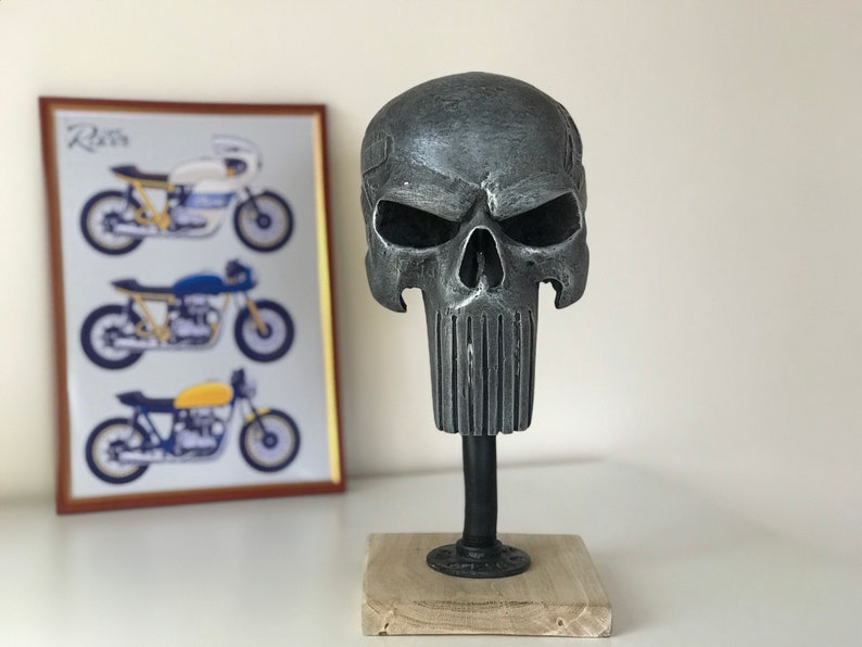 May include: A grey metal skull sculpture with a detailed face and a textured beard. The skull is mounted on a black metal stand with a wooden base.