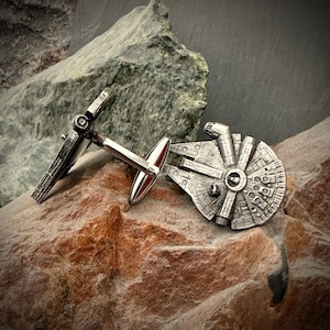 May include: Silver-toned cufflinks featuring a detailed Millennium Falcon design and a matching X-wing fighter. The cufflinks are set against a backdrop of textured brown and green rocks, creating a striking contrast.