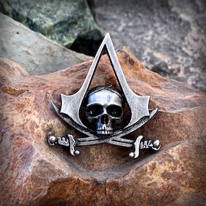 Puede incluir: Un pin de metal plateado con una calavera y huesos cruzados con un triángulo estilizado encima. La calavera lleva un pañuelo y los huesos cruzados son espadas.