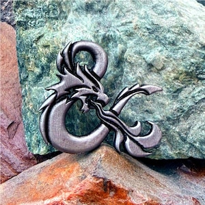 May include: A pewter-colored metal emblem featuring a dragon intertwined with an ampersand symbol. The dragon's head is detailed with scales and a fiery breath. The emblem is set against a backdrop of textured rocks.