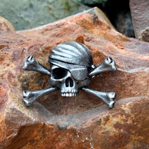 Può includere: Uno spillo in metallo argentato a forma di teschio di pirata e ossa incrociate, con un teschio che indossa una benda sull'occhio. Lo spillo è su una superficie di roccia marrone e ruvida.