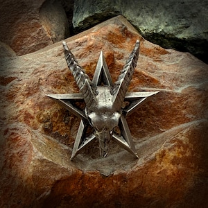 May include: A silver-colored pentagram pendant featuring a goat skull at its center. The pendant is set against a textured, reddish-brown stone background, creating a striking contrast. The horns of the goat are prominently displayed.