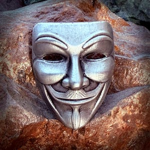 Peut inclure: Un masque argenté de Guy Fawkes avec un visage souriant, posé sur une surface rocheuse brune et texturée. Le masque présente une moustache, une barbiche et des sourcils arqués. Le masque est un symbole de protestation et d'anonymat.