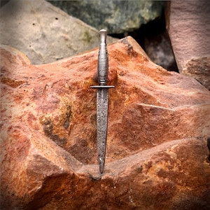 Könnte beinhalten: Ein silberfarbener Dolch mit einem strukturierten Griff und einer spitzen Spitze. Der Dolch liegt auf einem braunen, strukturierten Stein.