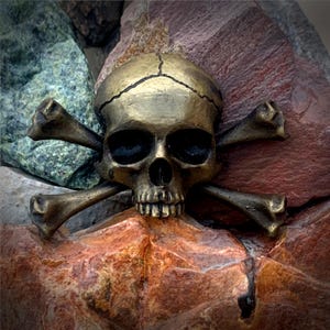 Puede incluir: Un diseño de calavera y huesos cruzados de color dorado. La calavera está detallada con una frente agrietada y una sonrisa de dientes. Los huesos cruzados son gruesos y tienen extremos redondeados.