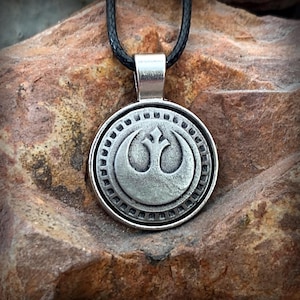 May include: A silver pendant necklace with the Rebel Alliance symbol from Star Wars. The circular pendant is suspended from a black cord. The pendant has a detailed design with a raised emblem and a textured border.