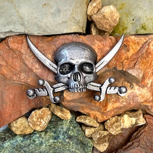 May include: A silver-colored skull and crossbones emblem. The skull is centered with two crossed swords behind it. The emblem is set against a background of various brown and green rocks.