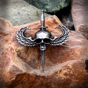 May include: A silver skull with wings and a dagger, a military-style pin.