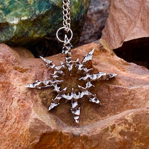 May include: A silver-toned pendant necklace featuring a starburst design with a textured, geometric pattern. The pendant is hanging from a silver chain.