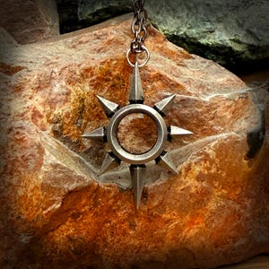 May include: A silver pendant necklace with a starburst design. The pendant has a central ring with eight pointed spikes around the edge. The necklace hangs from a chain. The pendant is displayed against a textured, reddish-brown rock.