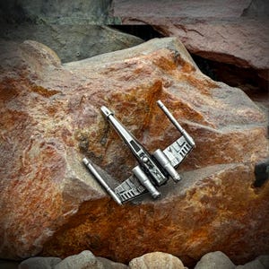 May include: A silver starfighter model with extended wings, positioned on a large, textured brown rock. The model has detailed features and is set against a natural background of rocks and stones.