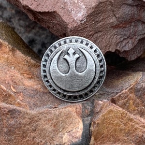 May include: A silver-toned metal pin featuring the Rebel Alliance symbol from Star Wars. The symbol is a white silhouette of a bird's wing with a circle around it.
