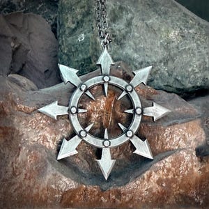 May include: A silver-colored pendant necklace featuring a stylized eight-pointed star design with arrow-shaped points. The pendant is suspended from a delicate chain. The necklace is displayed against a textured stone background.