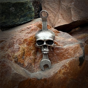 May include: A silver-colored pendant featuring a skull design integrated with a wrench. The skull has dark eye sockets and a detailed jawline. The wrench design includes both a closed and open end. The pendant is displayed against a textured, reddish-brown rock background.