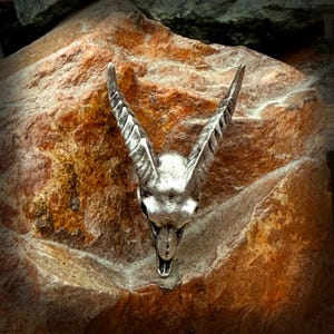 May include: A detailed silver sculpture of an animal skull with prominent, curved horns. The skull is set against a textured, reddish-brown stone background. The horns are a key feature, adding to the sculpture's dramatic appearance. The overall effect is striking.