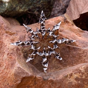 Puede incluir: Un colgante plateado en forma de estrella con un diseño geométrico texturizado. El colgante tiene una forma de estrella puntiaguda con un agujero central.