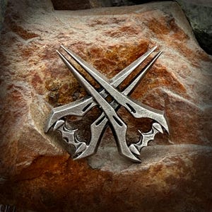 Op de afbeelding: Een zilverkleurig metalen embleem met twee gekruiste, gestileerde messen. De messen hebben ingewikkelde ontwerpen en zijn geplaatst tegen een getextureerde, bruine stenen achtergrond. Het embleem heeft een gotisch of fantasy thema.