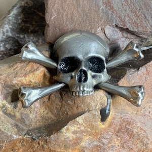 Op de afbeelding: Een zilveren schedel en gekruiste beenderen ontwerp. De schedel heeft zwarte oogkassen en een gedetailleerde mond. De gekruiste beenderen zijn gemaakt van twee zilveren beenderen die over elkaar gekruist zijn.