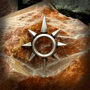 Puede incluir: Un diseño de estrella metálica plateada con ocho puntas afiladas, centrado alrededor de un anillo circular. La estrella está colocada sobre un fondo de piedra texturizada de color marrón rojizo.