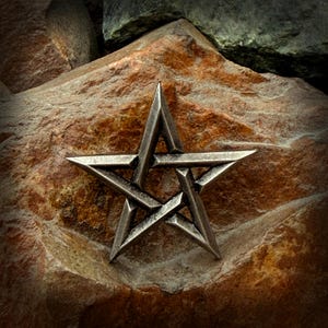 Può includere: Una stella pentagramma color argento, con punte affilate e angolari, poggia su una superficie di pietra marrone-rossastra strutturata. Il design della stella è dettagliato, con linee e bordi visibili. Lo sfondo presenta più pietra, creando un'estetica naturale e terrosa.
