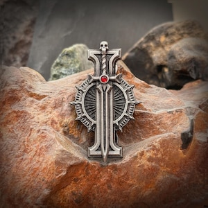 May include: A silver-colored pendant featuring a sword design with a red gem in the center. The pendant has a skull at the top and is set against a textured, reddish-brown stone background.