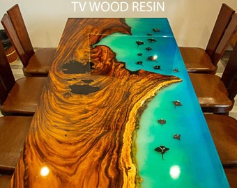Op de afbeelding: Een grote houten tafel met een inleg van turquoiseblauwe epoxyhars. De hars heeft een ontwerp van zwarte pijlstaartroggen die in het water zwemmen. De tafel is gelabeld met "TV WOOD RESIN".