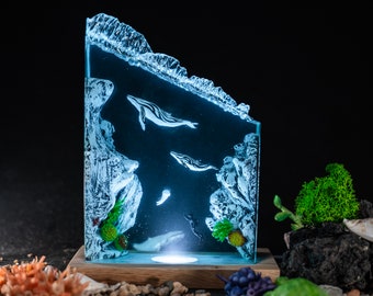 May include: An illuminated resin sculpture of an underwater scene, featuring whales, jellyfish, and coral. The artwork is encased in a clear, angled block, mounted on a wooden base. The whales are white with black stripes, and the scene is lit with a soft blue glow.