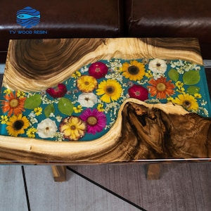 Puede incluir: Mesa de centro rectangular con estructura y patas de madera. La superficie de la mesa presenta una incrustación de resina transparente con flores de colores y un fondo turquesa. La madera tiene un patrón de vetas natural.
