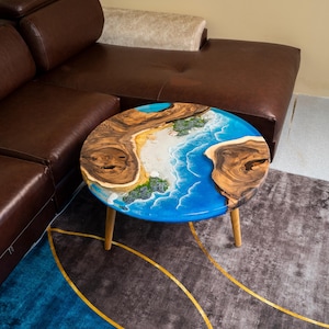 Puede incluir: Una mesa de centro redonda con un diseño único. La tapa de la mesa presenta una escena de playa con resina azul que representa el océano y secciones de vetas de madera. La mesa tiene tres patas de madera.