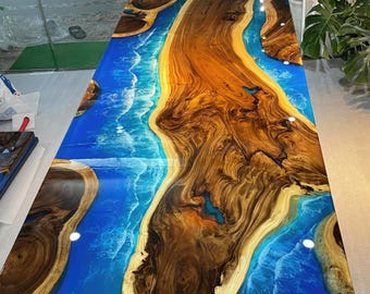 Op de afbeelding: Een grote houten tafel met een blauw en wit epoxyhars rivierontwerp. De tafel heeft een zwart metalen X-vormig onderstel.