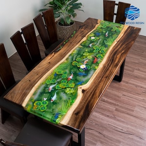 May include: A rectangular dining table featuring a live edge dark wood slab with a central river inlay made of green resin. The resin depicts a pond scene with koi fish, lily pads, and aquatic plants. The table is paired with six dark wood chairs.