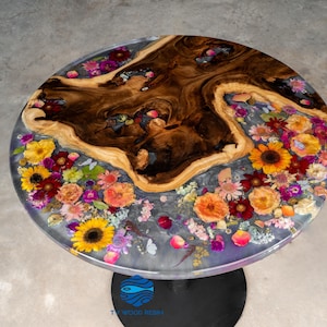 Puede incluir: Mesa redonda de madera con un centro de vetas de madera marrón oscuro y una parte superior de resina transparente. La resina está incrustada con flores coloridas, incluyendo girasoles y rosas. La mesa tiene una base de metal negro.