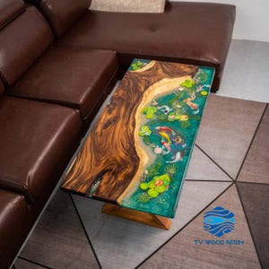Puede incluir: Mesa de centro rectangular con base de madera y tapa con diseño de estanque de carpas koi. La superficie de la mesa combina la veta natural de la madera con resina transparente de color turquesa, mostrando carpas koi y nenúfares. La mesa mide aproximadamente 122 cm de largo.