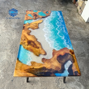 Könnte beinhalten: Rechteckiger Holztisch mit einem Harzdesign, das eine Strandszene darstellt. Der Tisch hat einen hellfarbigen Strand, türkisfarbenes Wasser und Holzakzente. Der Tisch wird von schwarzen Metallbeinen getragen.