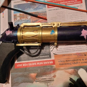 Puede incluir: Una pistola de juguete pintada a mano con un cañón dorado, un mango negro y detalles azules y rosas. La pistola está hecha de plástico y tiene un diseño realista.