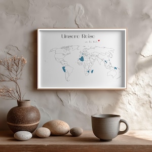 Könnte beinhalten: Gerahmter Kunstdruck einer Weltkarte mit dem Text "Unsere Reise am die Welt." Die Karte zeigt blau markierte Länder. Eine dekorative Vase, Steine und eine Tasse stehen auf einer Holzoberfläche.