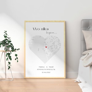 May include: Framed print featuring a heart-shaped map with a red heart marking a location. The text "Wo alles begann..." is at the top, with names and coordinates below. The frame is light-colored.