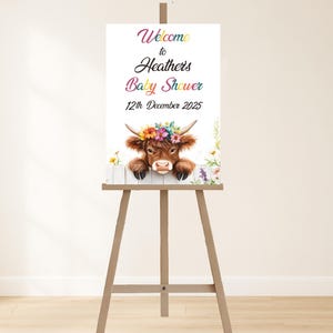 May include: A wooden easel supports a baby shower sign. The sign says "Welcome to Heathers Baby Shower 12th December 2025." A watercolour illustration of a Highland cow wearing a floral crown is featured above a white picket fence.