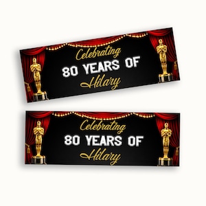 Może przedstawiać: Prostokątny baner z czarnym tłem, czerwonymi zasłonami po bokach i łańcuchami świetlnymi u góry. Tekst brzmi Celebrating 80 YEARS OF Hilary w białym i złotym kolorze. Dwie złote statuetki stoją na cokołach po obu stronach baneru.