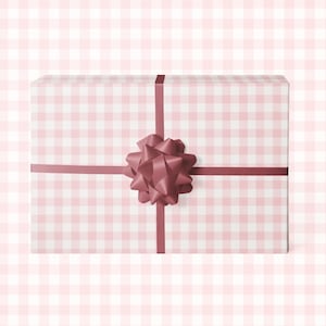 Puede incluir: Una caja de regalo rectangular envuelta en papel de cuadros rosa y blanco. Una cinta burdeos cruza la caja, asegurada con un lazo a juego. El fondo es un suave patr&oacute;n de cuadros rosas.