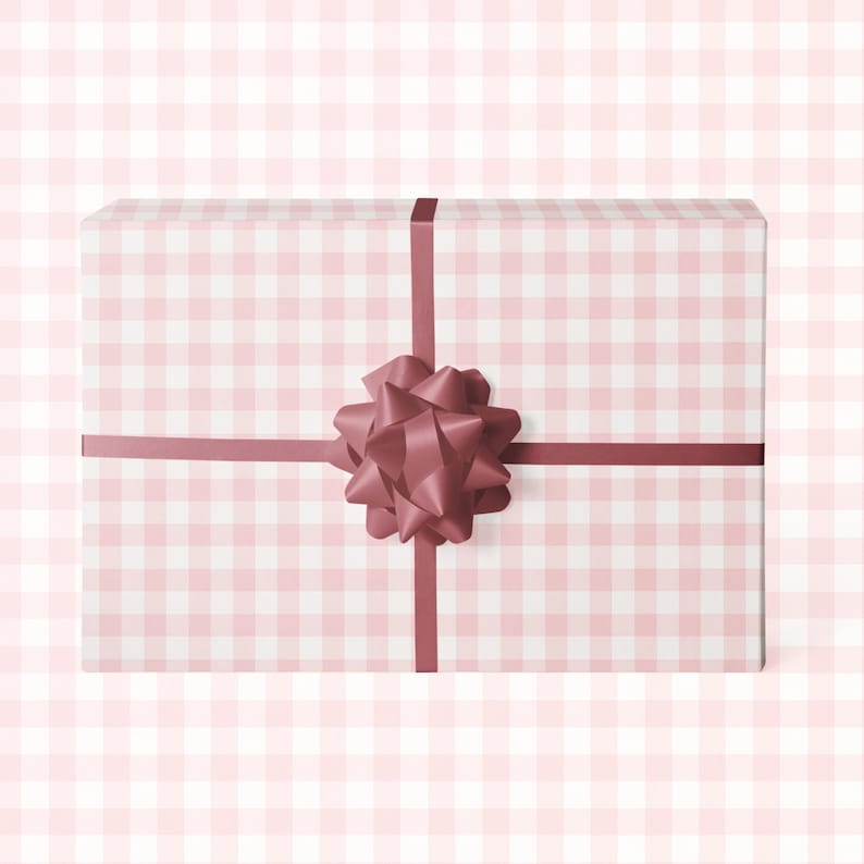 Puede incluir: Una caja de regalo rectangular envuelta en papel de cuadros rosa y blanco. Una cinta burdeos cruza la caja, asegurada con un lazo a juego. El fondo es un suave patr&oacute;n de cuadros rosas.