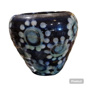May include: A handcrafted ceramic vase with a dark blue glaze and a pattern of white and light blue floral designs. The vase has a rounded shape and a textured surface, adding to its unique aesthetic.